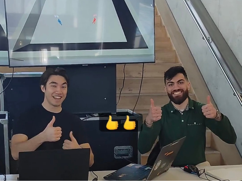 A picture of Zain and Marcus, with a TV behind them and game controllers on the table in front of them, holding their thumbs up.