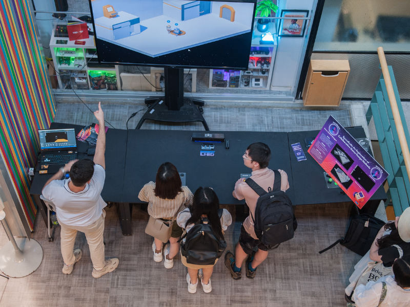 An aerial view of a table with built-in game controllers and a large TV behind it, multiple people gathered in front of it, one individual pointing at the screen.