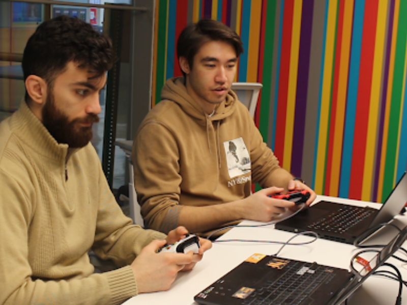 Zain and Marcus holding game controllers hooked up to laptops their focus on the screens in front of them. 