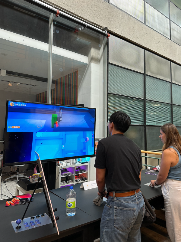 Two individuals are standing in front of a table with built-in game controllers and a large TV behind it, navigating the game at the MDM Industry Day showcase.