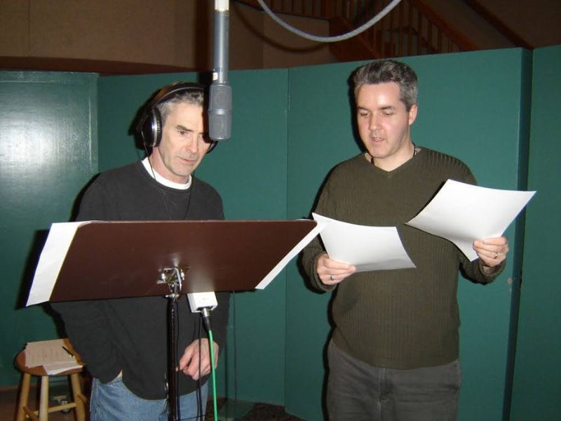  Two men standing in a recording booth holding pieces of paper.