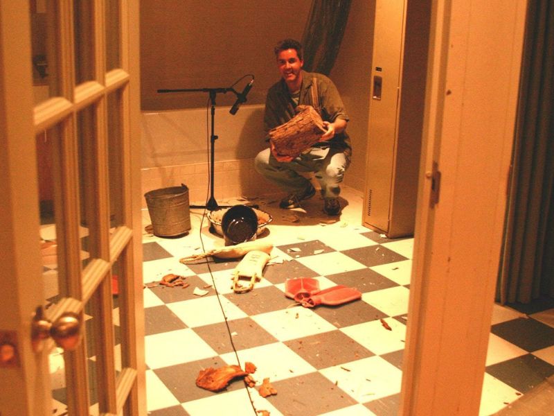  A man crouches in a corner holding a chopped piece of wood close to a microphone.