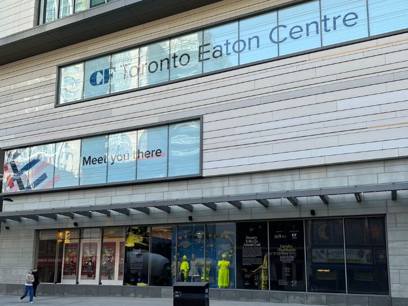 Exterior view of the Eaton Centre mall window displays showing faculty highlights poster and mannequins wearing neon yellow and grey outfits.