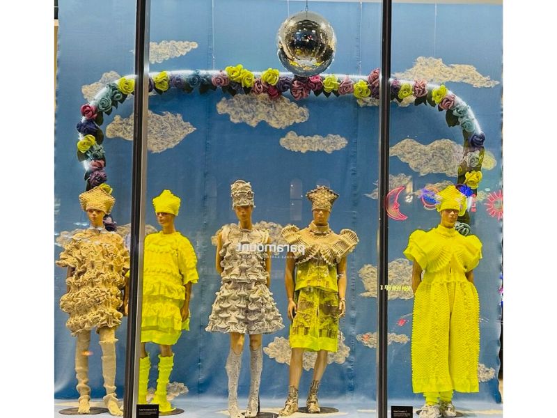 4 Mannequins displayed in the windows of the Eaton Centre. Mannequins are wearing neon yellow and muted grey outfits. The backdrop of the display is blue and white to resemble the sky and clouds. 