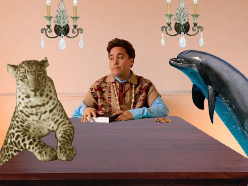 Chris E. Vargas sits at a table under two chandeliers, holding a book. A cheetah and dolphin are on either side of him.