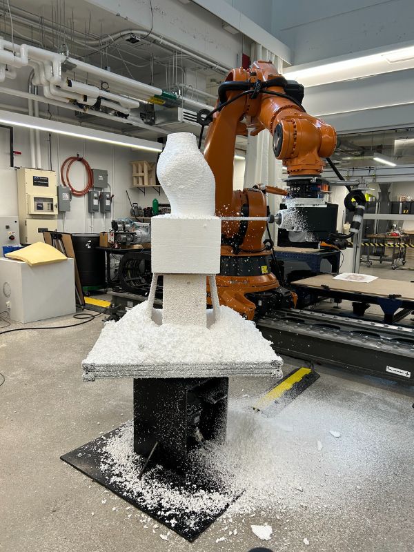 A Kuka robotic arm carves a 3D form out of white foam on a table in the Design and Technology Lab.