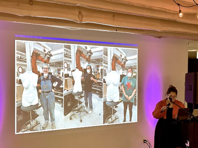Caron Phinney stands beside a slideshow showing images of three participants, each standing beside a white foam 3D model of their torso. Caron is talking into a microphone in front of a seated audience in The Catalyst.