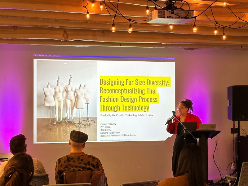 Caron Phinney stands beside a slideshow of her work titled Designing For Size Diversity: Reconceptualizing The Fashion Design Process Through Technology. She is talking into a microphone in front of a seated audience in The Catalyst.