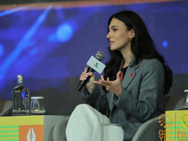 A woman wearing a gray blazer speaks into a microphone to a crowd outside of the frame of the image