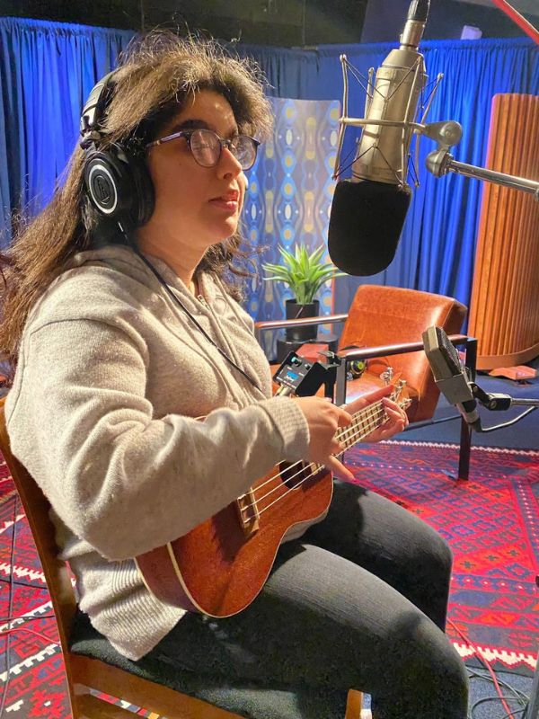 A women with long brown hair and an oversized grey sweater sitting down in a chair in a recording studio. She’s holding a ukulele while smiling happily.