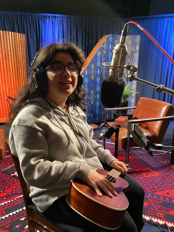  A women with long brown hair and an oversized grey sweater sitting down in a chair in a recording studio. She’s holding a ukulele while smiling happily