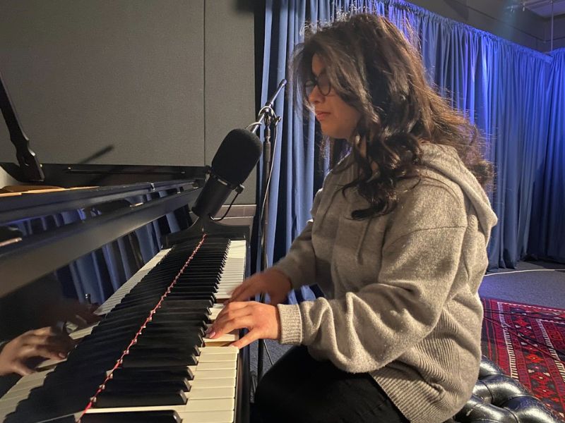 A women with long brown hair and an oversized grey sweater sitting down in a chair in a recording studio. She’s playing the piano and singing into a microphone resting on top of the piano.