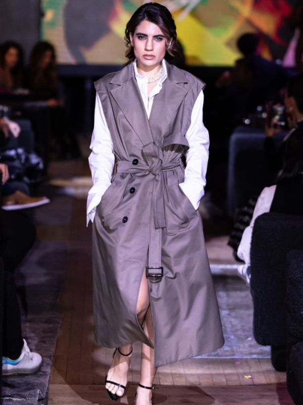 Model staring into the camera while walking down the runway wearing a white blouse and sleevless grey trenchcoat. 