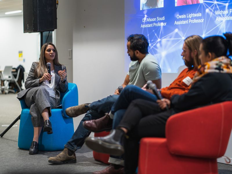 4-person panel discussion showing the moderator asking a question to the 3 panelists while holding a microphone and sitting on red and blue couches