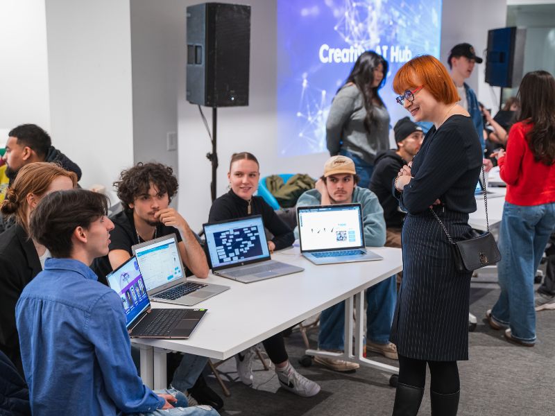 Student showcase showing people mingling and conversing with eachother. A group of students presenting their work via laptops and addressing questions made by audience memebers