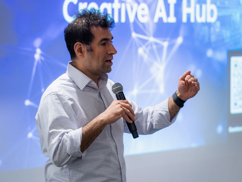 A man wearing a white dress shirt and speaking into the microphone while giving a presentation with a projector screen showcasing "Creative AI Hub" in the background