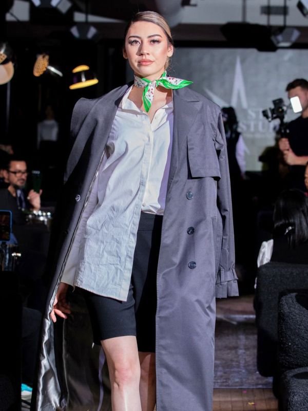 Woman model walking down fashion runway wearing a white blouse, black shorts, and grey trench coat. A green and pink bandana is tied around her neck. In the background, many onlookers are sitting or taking photos of the model