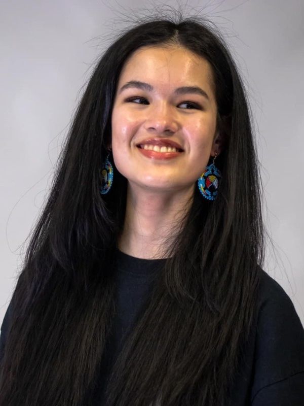 Woman smiling with dark brown hair wearing a black shirt and turqoise earrings with the Indigenous medicine wheel on it. 