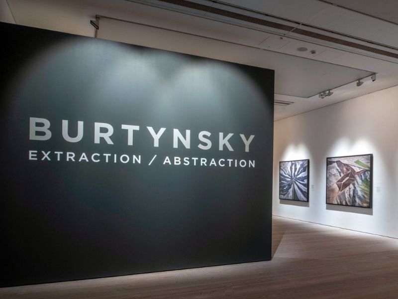 Art gallery exhibition with a black wall titled "Burtynsky Extraction/ Abstraction" at the forefront of the gallery entrance. Art pieces can be seen at the far right of the room. 