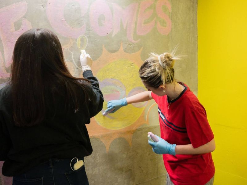Two students use colorful chalk to draw a sun on a concrete wall.
