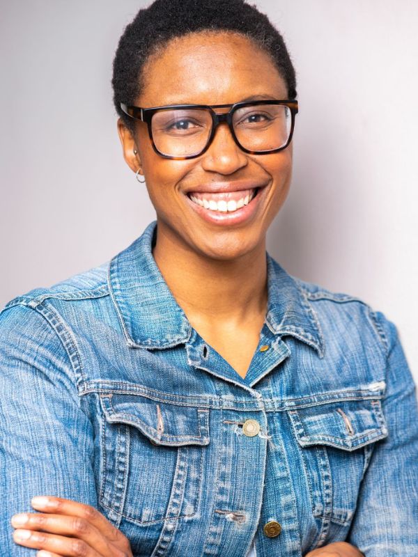 Woman wearing a jean jacket and black glasses with arms crossed smiling for a photo