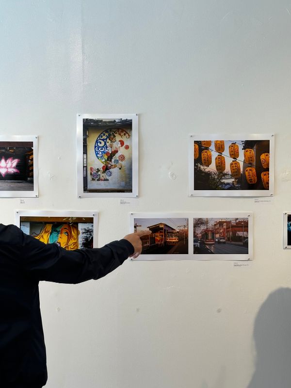 A man halfway out of the frame points at a wall with a picture of two train stations.
