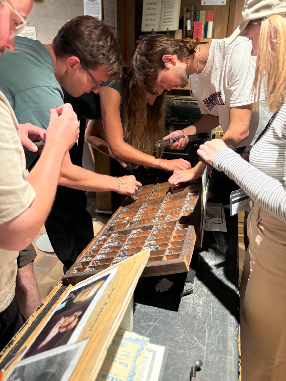 Detailed medium-long shot of five trainees observing the shop’s letterpress trays.
