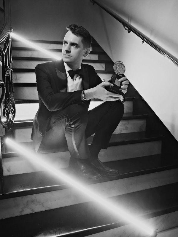 A black and white image of a man in a tuxedo sitting on a set of stairs holding an award