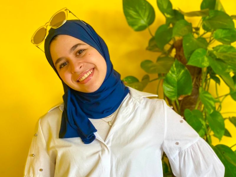 Portrait of Dina Badawi standing in front of a yellow wall and a green plant 