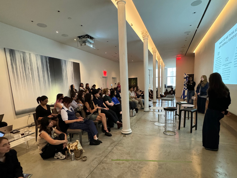Three presenters stand in front of a slide deck with students sitting down, listening to them. 