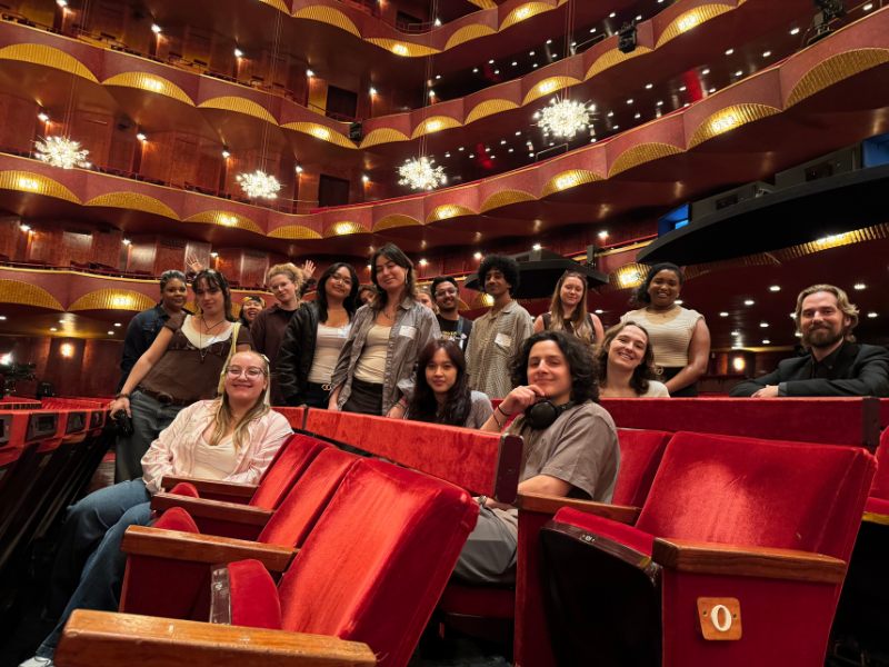 Students sitting down on red theatre seats, smiling at the camera.