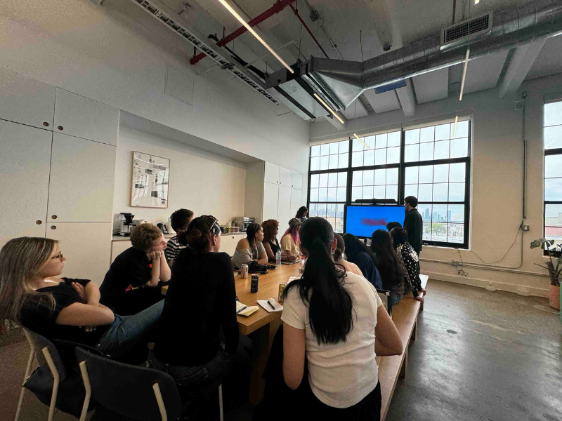 Students are gathered around a boardroom table, looking at a TV. Standing next to the TV is a presenter. 