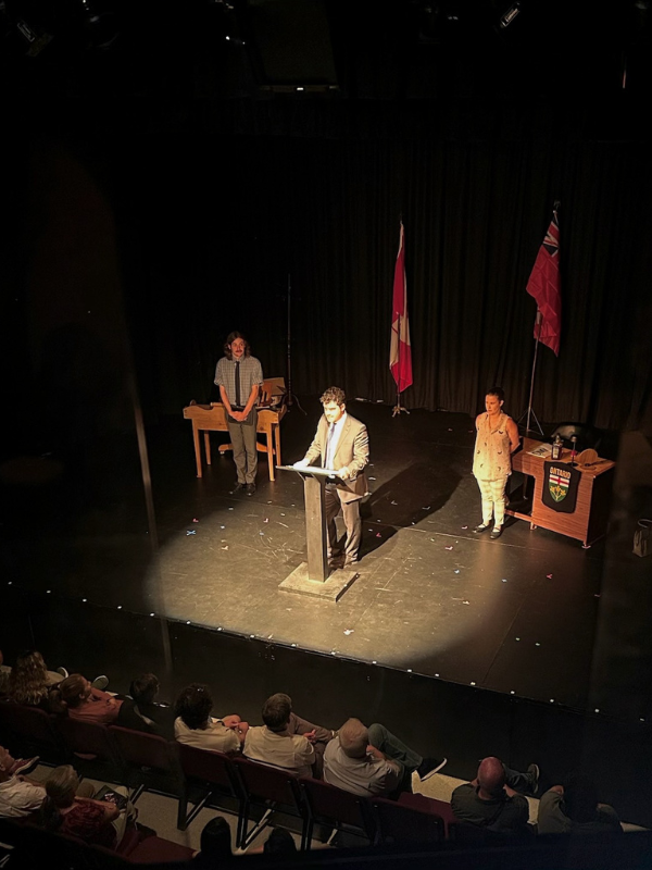 Overhead shot of three actors on stage in a dark theatre, with a spotlight on the main actor and the audience visible at the bottom.