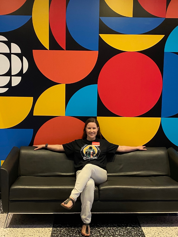 Wide shot of Victoria Sullivan sitting on a couch and smiling at the camera at a CBC location. The couch is black, and the wallpaper shows loud shapes that are red, orange, yellow, and blue.