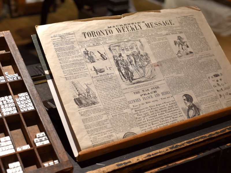 A full shot of a half-folded newspaper propped up on a podium. The title of the newspaper reads “Mackenzie’s Toronto Weekly Message” and features other smaller articles and images throughout.
