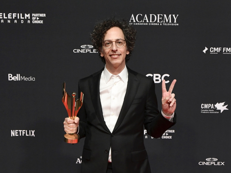 A person in a black suit stands on the red carpet holding an award in one hand and making a peace sign with the other in front of a black backdrop with logos.