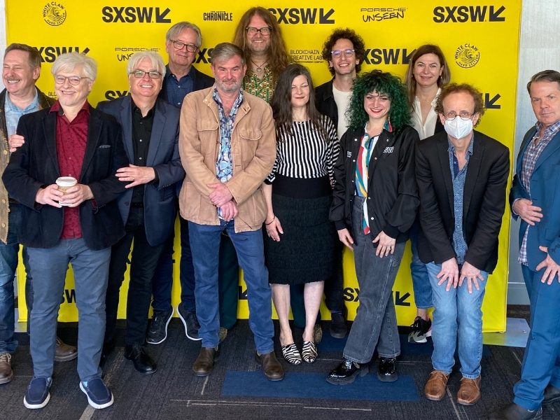 A group of thirteen people stands together smiling in front of a bright yellow SXSW backdrop.