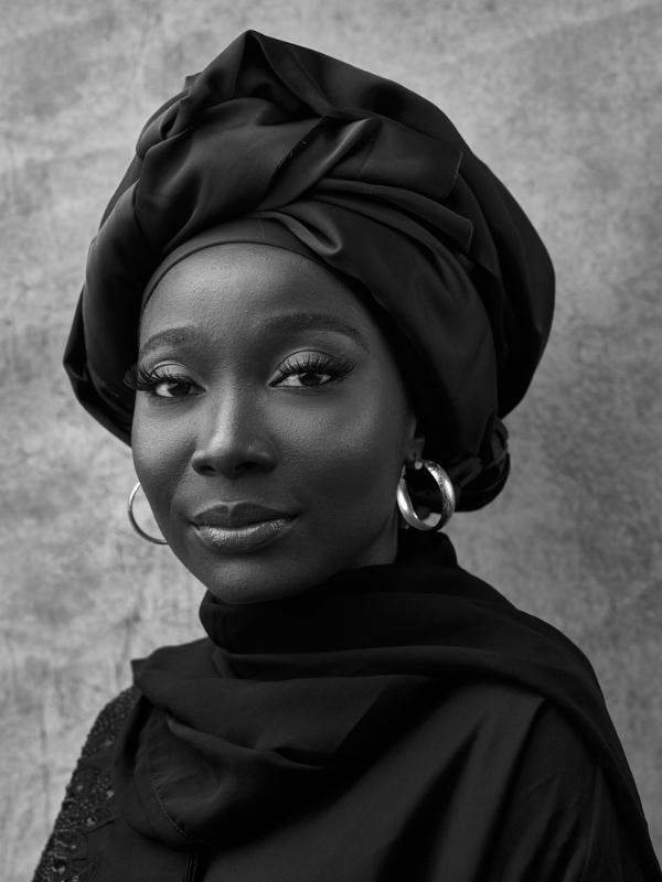 Black-and-white portrait of Azizah Adunola Alawusa wearing a headwrap and hoop earrings, looking toward the camera.
