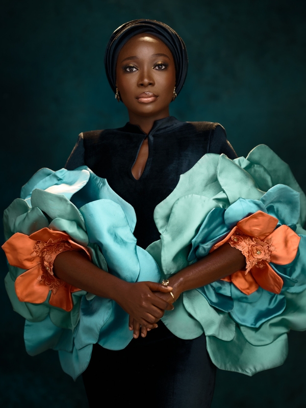 Azizah Adunola Alawusa poses in a dark outfit with large teal-and-orange floral sleeves against a deep green background.