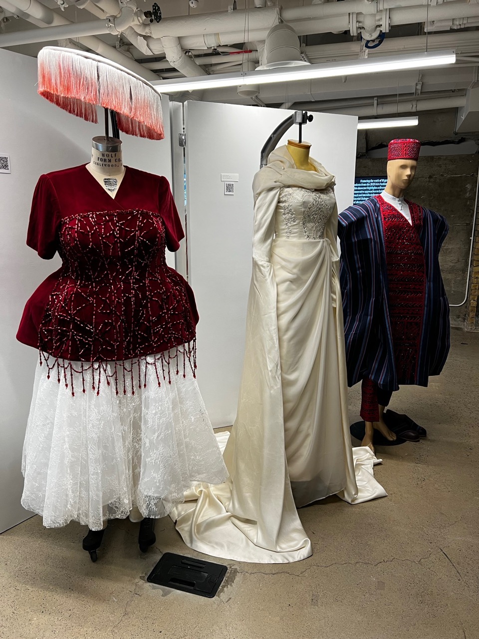 Three mannequins display garments designed by Azizah Adunola Alawusa, including a red beaded dress, a white gown, and a traditional striped outfit.