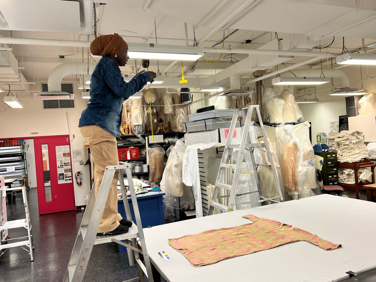 Azizah Adunola Alawusa stands on a ladder photographing a garment in TMU’s fashion studio filled with materials and dress forms.