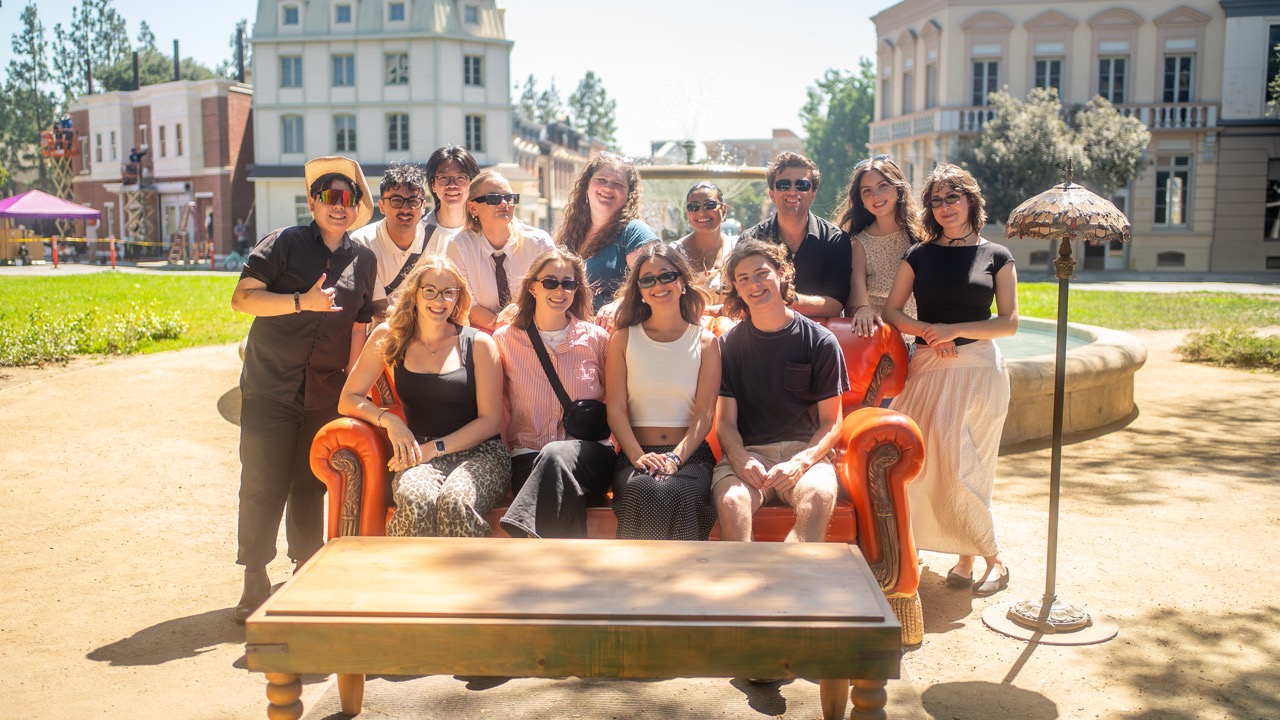 Alt text: Thirteen participants of RTA in LA posing for a photo outdoors on a replica of the orange couch from the TV show Friends. They are sitting and standing around the couch. The setting is sunny with classic-style buildings in the background, a fountain behind them, and a wooden coffee table in the foreground.