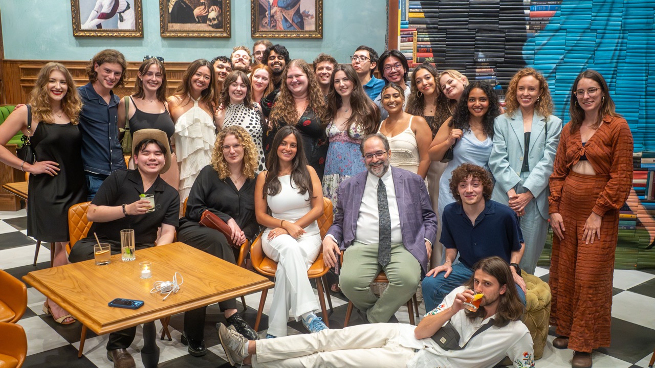 A group of students and their professor, all participants in the RTA program in LA, are together in a lively indoor setting. Some are standing, while others sit, all dressed in a mix of formal and casual attire. The background features colourful walls and bookshelves.