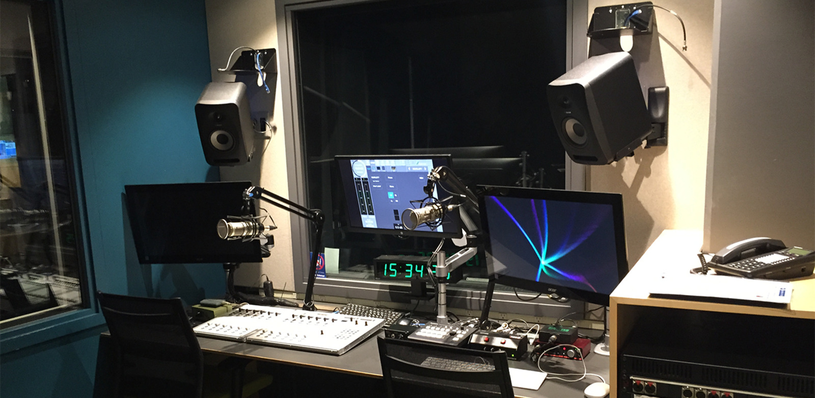 A modern radio studio with two microphones on adjustable arms, two computer monitors, a mixing console, speakers mounted on the wall, and a digital clock.