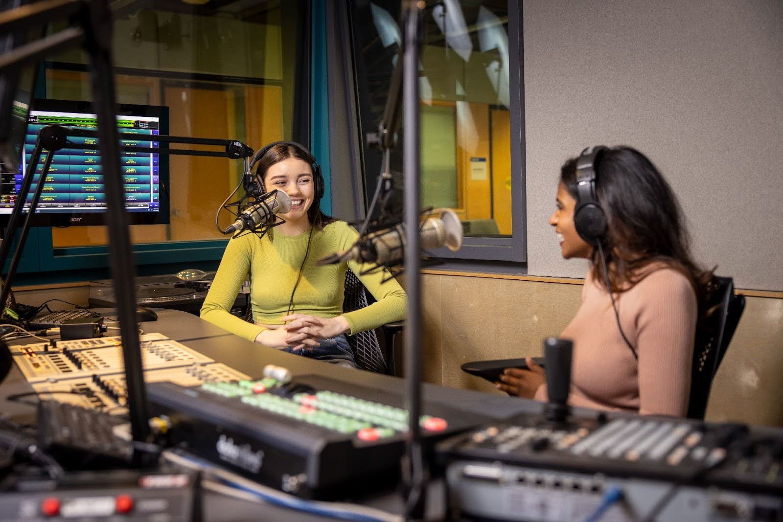 Two women are sitting at the Allan Slaight Radio Institute in front of microphones and switchboards. They are looking at each other and appear to be in the middle of a conversation."