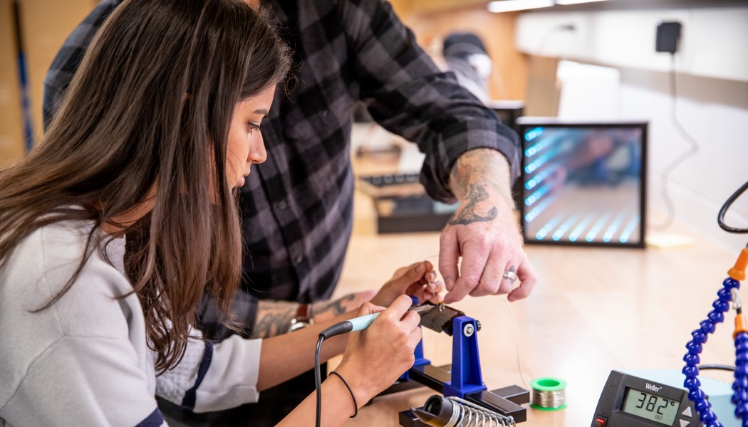 A student is guided by an instructor while soldering components for a technology project.