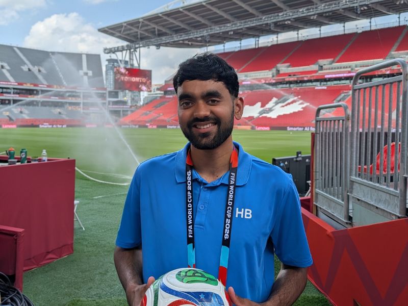 Ali Imam smiles while standing beside the pitch at Toronto Stadium during the FIFA World Cup 2026. Wearing a blue FIFA Host Broadcast (HB) Intern Programme polo shirt and accreditation, he holds a FIFA match ball with the stadium, soccer field and sprinklers visible in the background.