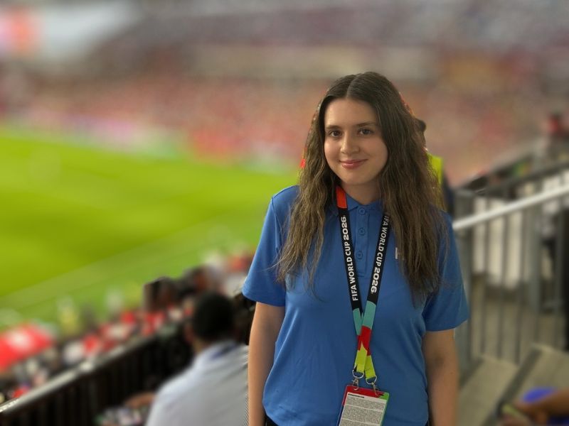 Cristina Carbonelli stands at a broadcast position overlooking the pitch at Toronto Stadium during a FIFA World Cup 2026 match. Wearing a blue FIFA Host Broadcast (HB) Intern Programme shirt and accreditation, she poses with the field and a packed stadium visible behind her.