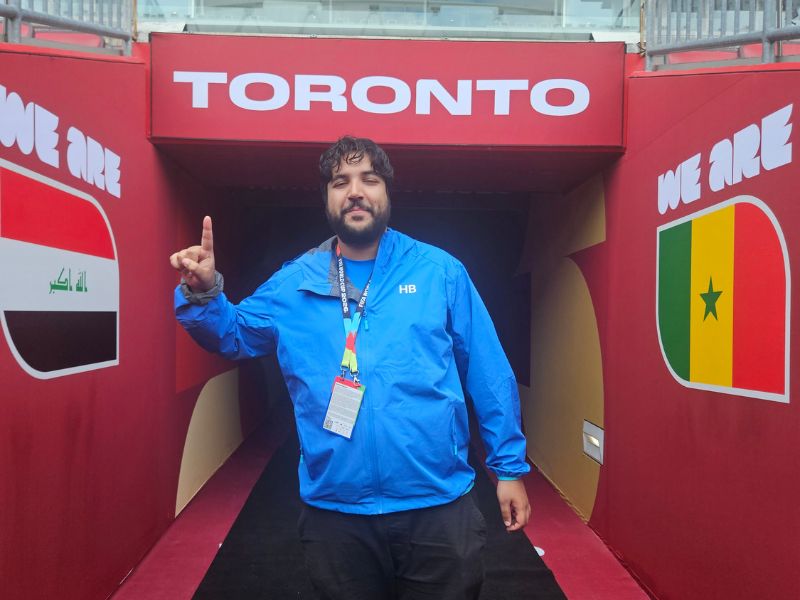Harsh Kumar stands in the player tunnel at Toronto Stadium beneath a large "TORONTO" sign during the FIFA World Cup 2026. Wearing a blue FIFA Host Broadcast (HB) Intern Programme jacket and accreditation, he poses between tournament-branded walls displaying national team graphics.