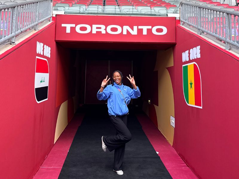 Jessica Edet-Nankere smiles while posing in the player tunnel at Toronto Stadium during the FIFA World Cup 2026. Wearing a blue FIFA Host Broadcast (HB) Intern Programme jacket, she stands beneath a large "TORONTO" sign with tournament-branded walls on either side of the tunnel.